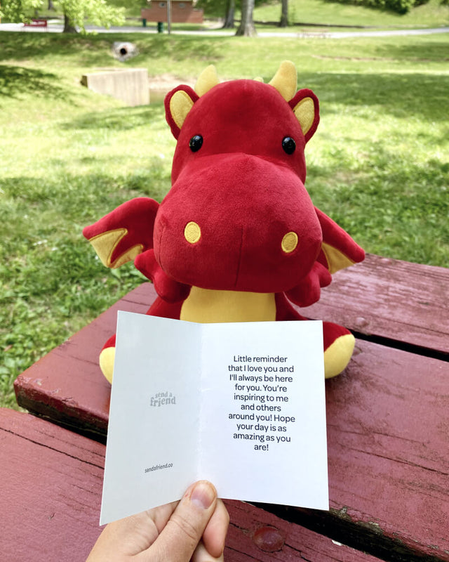 Photo of a hand holding a note card with red and yellow Duke the Dragon plushie in the background sitting on a picnic table outdoors