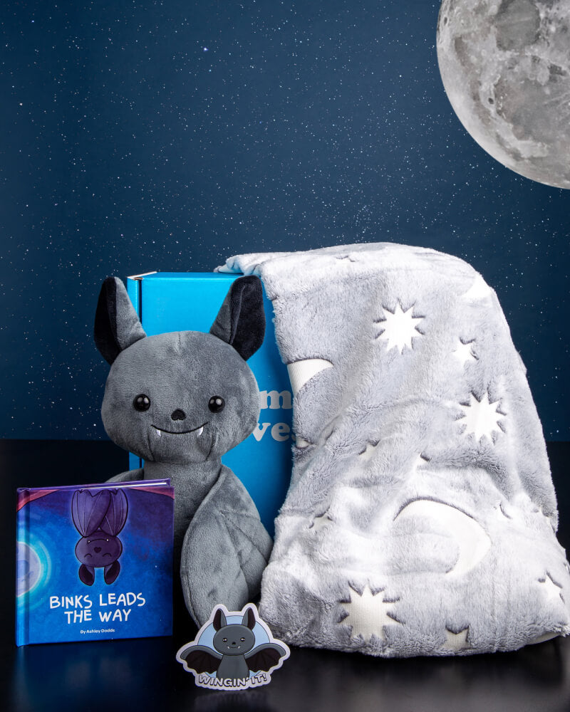 Photo of Binks the Bat plushie with his storybook, bat sticker, moon and stars gray blanket, and blue box with the moon and stars on a blue background