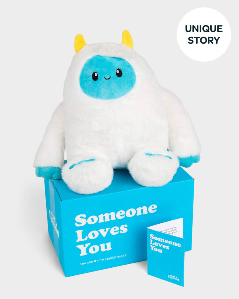 White yeti plushie with blue face and yellow horns sitting on top of SendAFriend box and next to notecard both in signature blue color