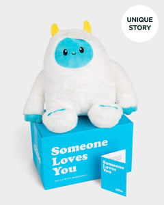 White yeti plushie with blue face and yellow horns sitting on top of SendAFriend box and next to notecard both in signature blue color