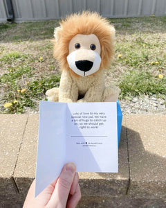 Photo of hand holding note card with tan Leroy the Lion plushie sitting in the background