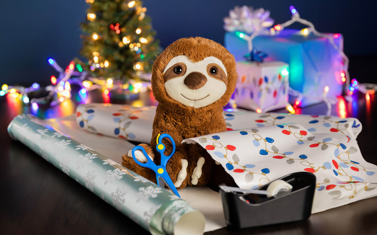 An image of Sam the Sloth with wrapping paper, scissors, tape. In the background is a present, Christmas lights, and a Christmas tree