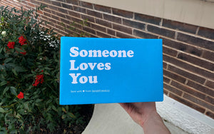 An image of a Someone Loves You box being held in front of a brick wall 