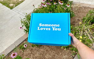 An image of a hand holding the signature "Someone Loves You" box with plants and flowers in the background