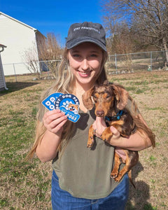 A smiling woman standing outside and wearing a baseball cap and holding three stickers and a small dog