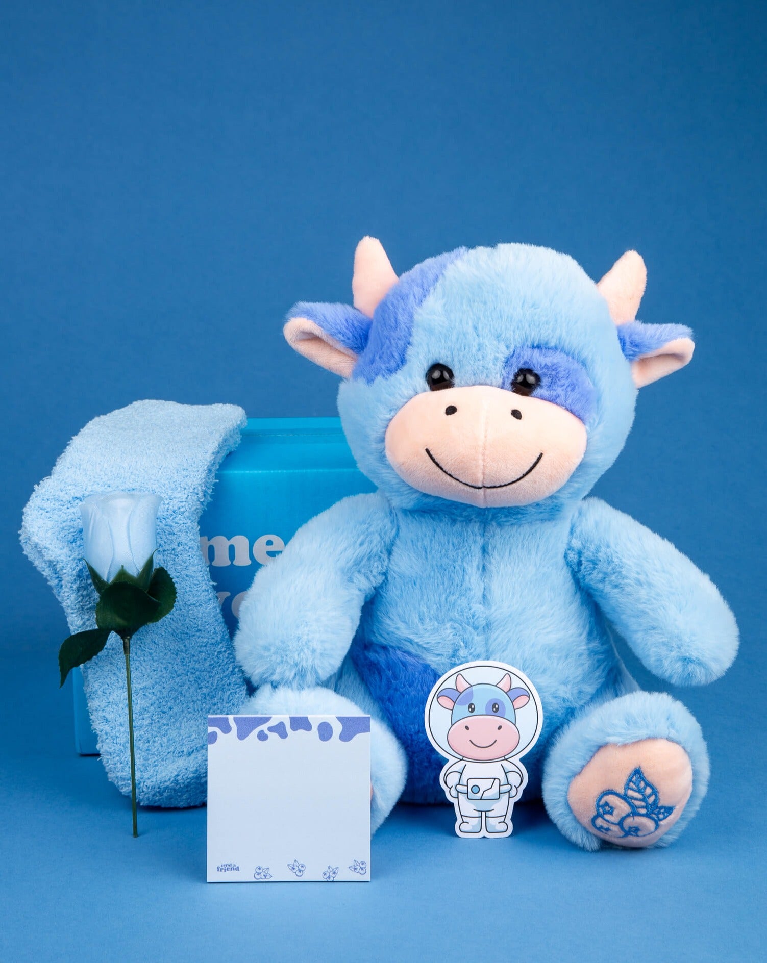 Photo of Beau the Blueberry Cow plushie with blue fuzzy socks, light blue silk rose, blueberry-themed sticky notes, blueberry cow sticker, and blue box on a blue background
