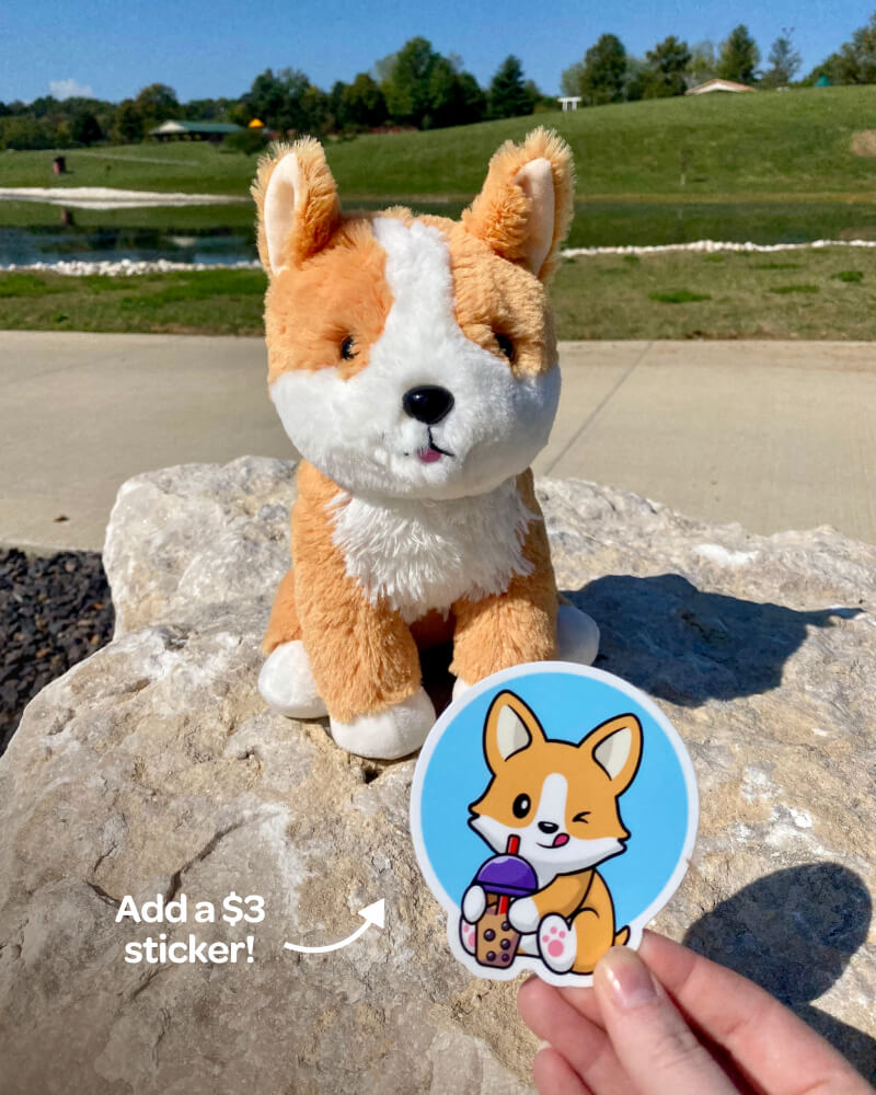 Photo of brown and white Comet the Corgi plushie outdoors sitting on a rock and a hand holding matching sticker available for additional $3