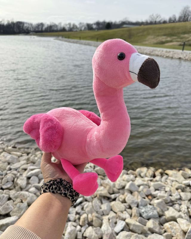 Side view photo of pink Faye the Flamingo being held up by a hand outdoors near a body of water