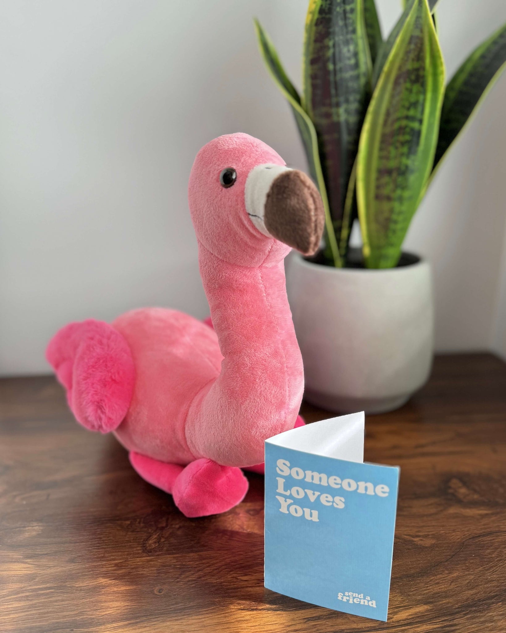 Photo of pink Faye the Flamingo sitting on a wood table with note card and a plant