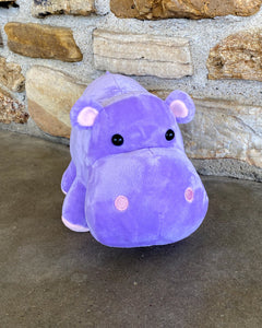 Photo of purple Harper the Hippo plushie sitting on concrete floor with rock wall in background 