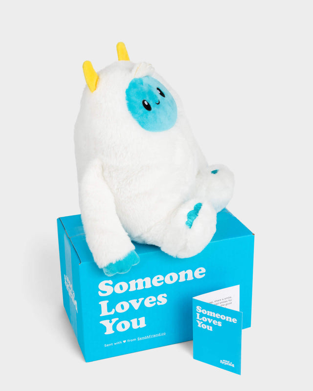 Side view photo of white Yuka the Yeti with blue face and yellow horns sitting on Someone Loves You box, and note card