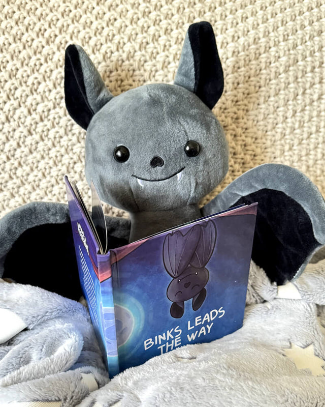Photo of Binks the Bat plushie with his open story book on a cream background