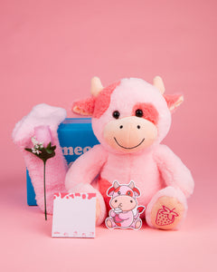 Photo of Sally the Strawberry Cow plushie with pink fuzzy socks, pink silk rose, strawberry-themed sticky notes, strawberry cow sticker, and blue box on a pink background