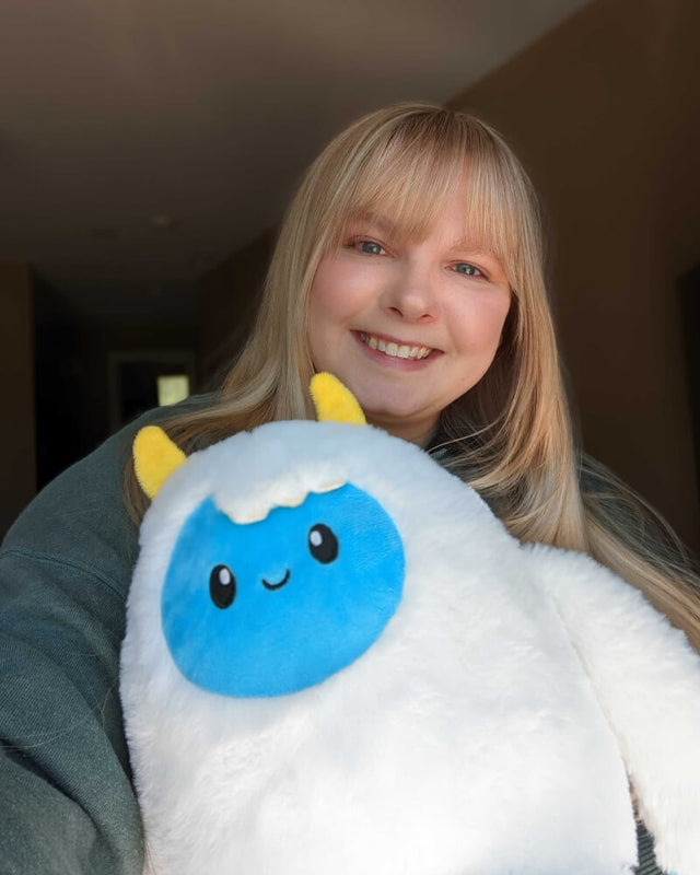 Photo of person smiling while holding white Yuka the Yeti plushie with blue face and yellow horns