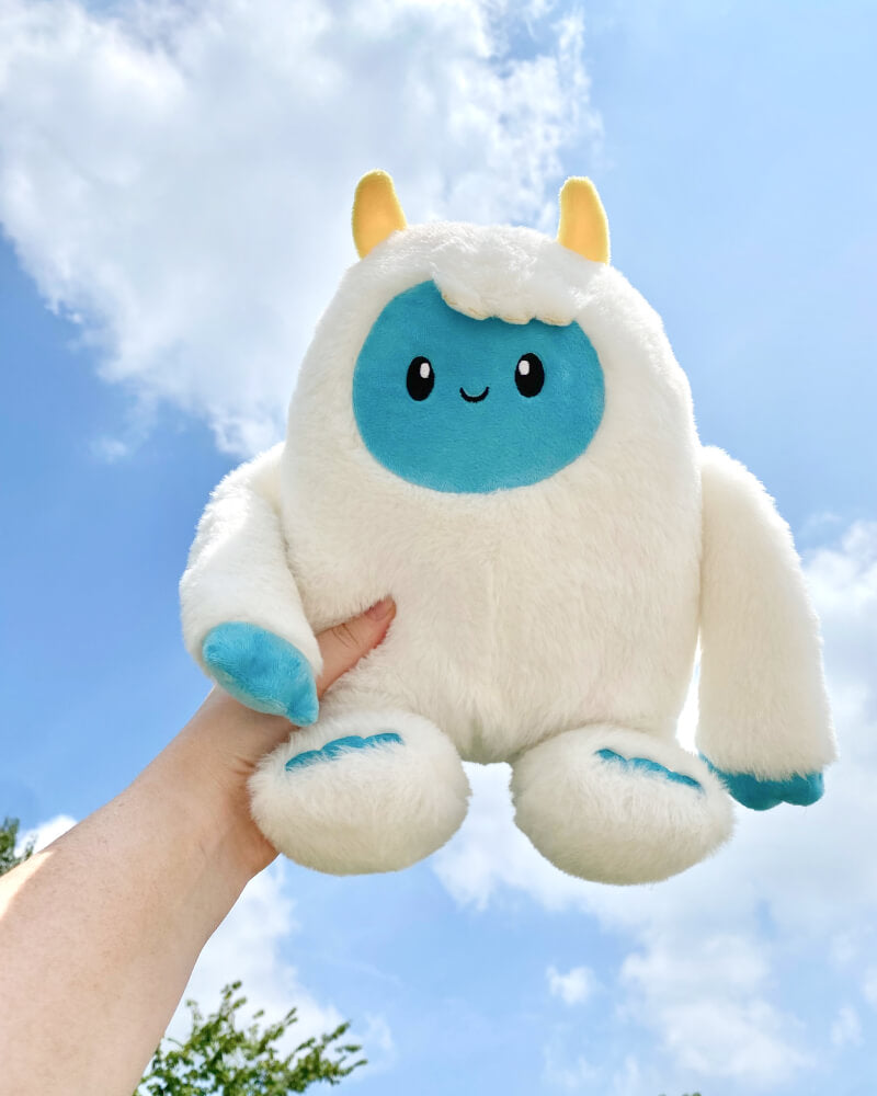 Photo of hand holding white Yuka the Yeti plushie with blue face and yellow horns, sky in the background