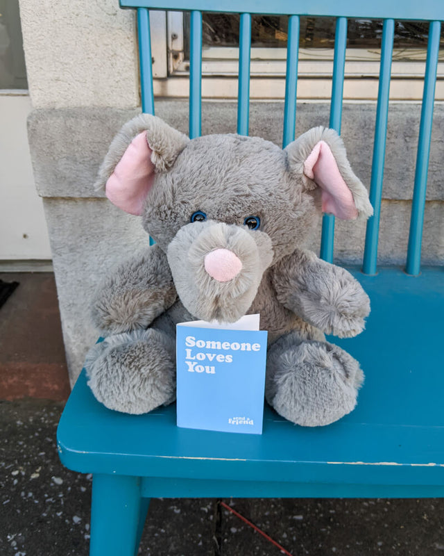 Photo of grey Eli the Elephant plushie sitting on blue chair with note card 