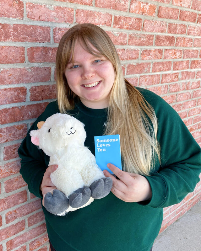 Photo of person standing next to brick wall holding white Gus the Goat plushie and note card