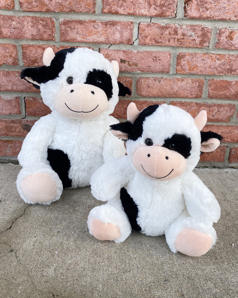 Photo of 14 inch and 10 inch black and white Cooper the Cow plushies sitting side by side with brick wall in the background