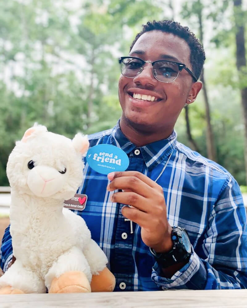 Photo of person outdoors holding up blue oval shaped SendAFriend tag attached to white Lawrence the Llama plushie