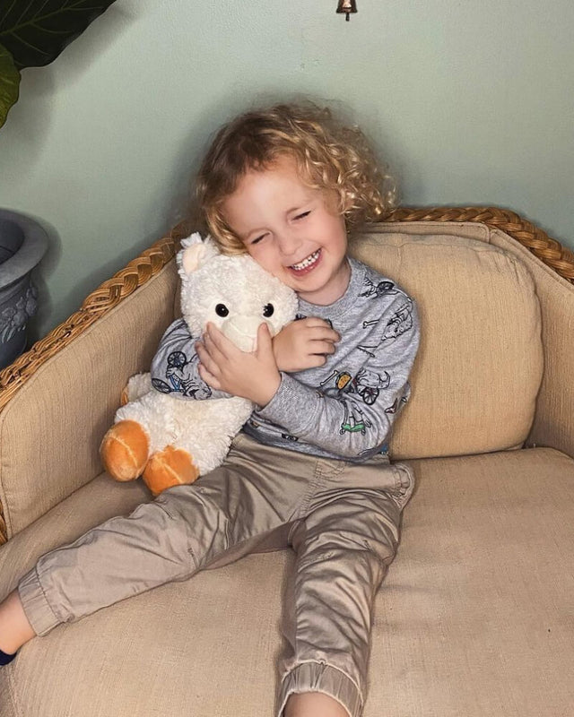 Photo of an adolescent sitting in a chair smiling while hugging white Lawrence the Llama plushie
