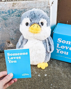 Photo of hand holding note card with grey and white Pepper the Penguin plushie sitting behind next to Someone Loves You box
