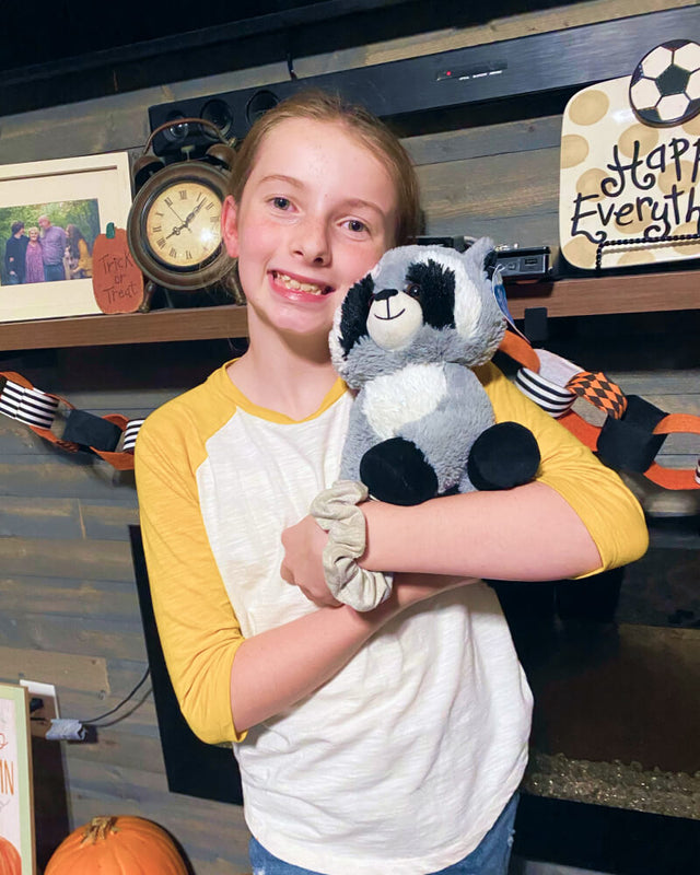 Photo of adolescent smiling while holding grey, black, and white Rosie the Raccoon plushie