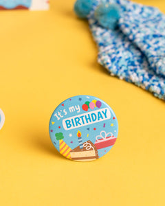 Photo of blue "It's my Birthday" pin featuring confetti, balloons, party hat, cake with candle, and present 