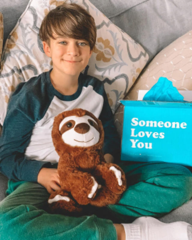 Photo of adolescent grinning while sitting and holding brown Sam the Sloth plushie next to Someone Loves You box