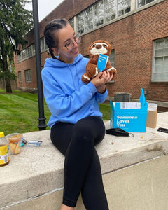 Photo of person smiling, sitting outdoors next to open Someone Loves You box, smiling while holding Sam the Sloth plushie and note card