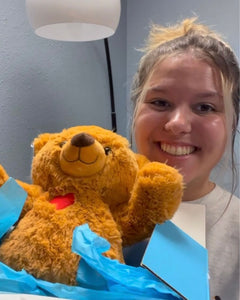 Photo of person smiling holding open box with brown Toby the Teddy Bear plushie in it