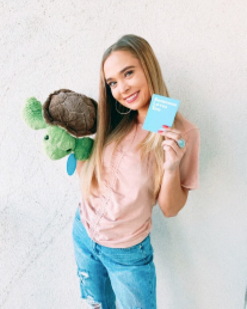 Photo of person smiling while holding green Tucker the Turtle with brown shell plushie and note card